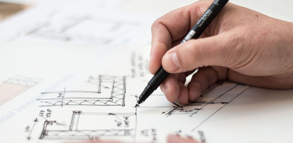 Desktop covered in design papers with hand of a man with a fine tip pen